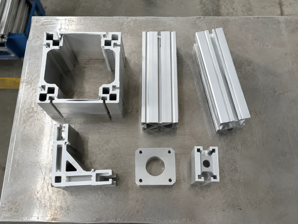 Six separate aluminum profile and plate samples are arranged on a metal worktable in an overhead view.